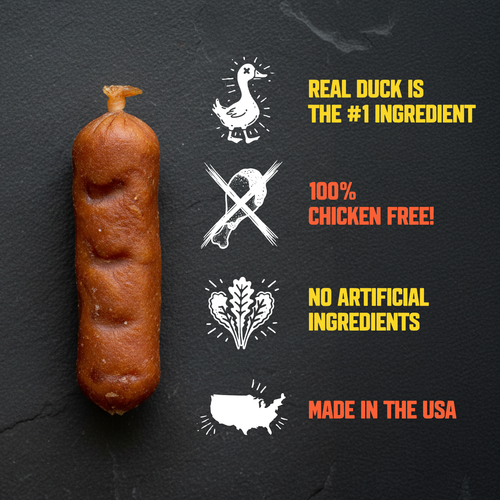 A single browned sausage or sausage-like food item stands on a dark textured background beside marketing callouts and icons: a duck illustration with 'REAL DUCK IS THE #1 INGREDIENT,' a crossed-out chicken symbol with '100% CHICKEN FREE!,' a heart-shaped vegetable icon with 'NO ARTIFICIAL INGREDIENTS,' and a U.S.